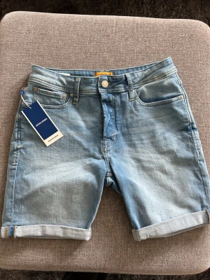 Ljusblå jeansshorts från Jack & Jones - Säljer ett par ljusblå jeansshorts från Jack & Jones med klassisk femficksdesign och uppvikta benslut. Shortsen har normal passform och stängs med dragkedja och knapp. Perfekta för varma dagar och enkel att matcha med olika stilar.
