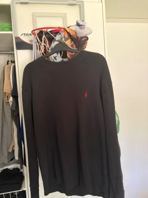 Mörkgrå stickad tröja från Polo Ralph Lauren - Mörkgrå stickad tröja från Polo Ralph Lauren med klassisk röd broderad logga på bröstet. Tröjan har rund halsringning och långa ärmar. Materialet är ribbstickat och ger en avslappnad men stilren look.