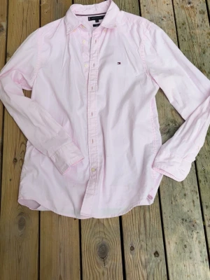Rosa skjorta slimfit från Tommy Hilfiger - Säljer en rosa skjorta från Tommy Hilfiger med klassisk krage och knappstängning framtill. Skjortan har lång ärm och en liten broderad logga på bröstet. Tillverkad i mjukt bomullstyg och har en avslappnad passform.