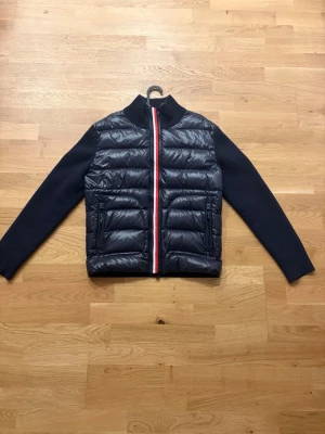 Moncler Hybrid Knitted Jacka - Snygg mörkblå dunväst med quiltat mönster och stickade ärmar. Dragkedjan framtill har en röd, vit och blå rand som ger en sportig touch. Hög ribbad krage och två stora fickor framtill. Perfekt för lager-på-lager och streetstyle.