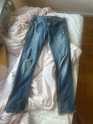 Blå jeans med grön dragkedja - Säljer ett par blå jeans med raka/utsvängda ben och slitna detaljer. Baksidan har en unik grön dragkedja på ena fickan och klassiska sömmar. Jeansen har en cool streetstil och är tillverkade i denim med en snygg tvätt.