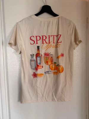 Beige t-shirt med Spritz-tryck - Säljer en beige t-shirt med färgglatt Spritz-tryck på både fram- och baksidan. Framsidan har en liten detalj med text och apelsin, medan baksidan har ett stort motiv med flaskor, glas, apelsiner och blommor. T-shirten har rund hals och korta ärmar.