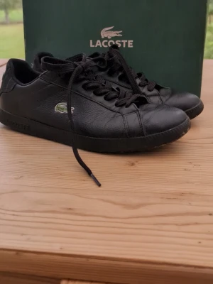 Svarta Lacoste sneakers i läder - Snygga svarta sneakers från Lacoste i läder med klassisk låg profil och grönt krokodilemblem på sidan. Skorna har svarta skosnören och diskret Lacoste-logga på sulan. Perfekta för en clean och stilren look.