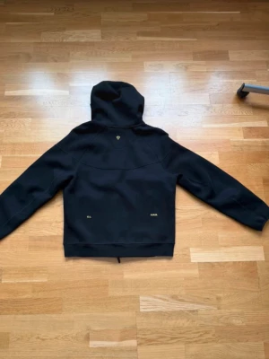 Svart hoodie från NOCTA med gul text - Snygg svart hoodie från NOCTA med gul text och detaljer. Hoodien har en klassisk passform med huva och ribbade muddar. Materialet ser ut att vara mjukt och bekvämt, perfekt för en streetwear-look. Gul NOCTA-logga på ärmen och små detaljer på ryggen.