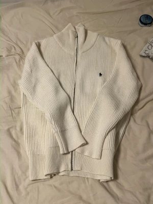 Vit stickad ziptröja från Polo Ralph Lauren - Klassisk vit stickad ziptröja från Polo Ralph Lauren med ribbad struktur och hög krage. Tröjan har dragkedja framtill och den ikoniska broderade loggan på bröstet. Perfekt för lager på lager och enkel att matcha till olika outfits.