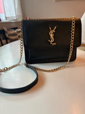 Yves Saint Laurent - Snygg svart axelväska  i slätt läder med guldfärgad kedja och ikoniska loggan framtill. Väskan har magnetlås, flera fack inuti och röd insida. Perfekt för dig som vill ha en klassisk och lyxig accessoar.