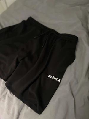 Svarta shorts från McKenzie - Svarta shorts från McKenzie med enkel design och vit logga på vänster ben. Shortsen har elastisk midja med snörning för skön passform. Perfekta för sommaren eller träning och tillverkade i mjukt syntetmaterial.