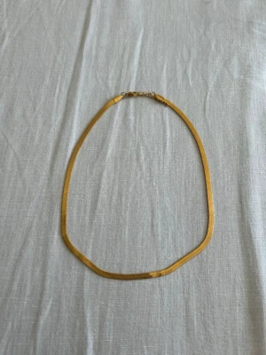 Guldigt platt halsband - Snyggt och stilrent halsband i guldfärgad metall med platt, ormliknande kedja. Halsbandet har en enkel låsning och passar perfekt för att ge en elegant touch till din outfit. Kedjan ligger platt mot huden och reflekterar ljuset snyggt.
