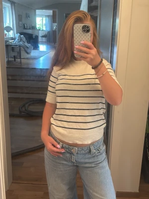 Vit randig kortärmad topp - Vit topp med mörkblå horisontella ränder och korta ärmar. Modellen är croppad och har en klassisk, enkel design som passar till jeans eller kjol. Tillverkad i mjukt material, perfekt för varmare dagar.