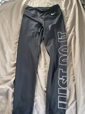 Svarta Nike leggings med tryck - Snygga svarta leggings från ” DRI- FIT” med vitt 'JUST DO IT'-tryck längs ena benet och liten Nike-logga vid midjan. Tight passform och stretchigt material som passar perfekt till träning eller chill. Hög midja för extra komfort.