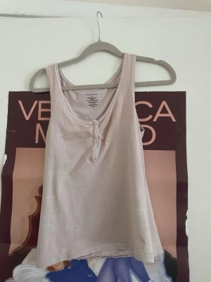 Ljusbeige ribbad tanktop med knappar - Säljer en ljusbeige ribbad tanktop med smala axelband och knappdetaljer framtill. Tillverkad i mjuk bomull med lite stretch för extra komfort. Perfekt till sommaren eller som lager på lager-plagg.