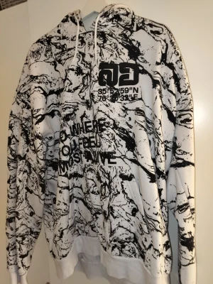 Vit och svart hoodie med marmorprint - Säljer en vit hoodie med svart marmorliknande print och textdetaljer på bröstet. Hoodien har huva med vita snören och ribbade muddar vid ärmslut och nederkant. Trycket inkluderar koordinater och texten 'WHERE YOU FEEL MOST ALIVE'. Perfekt för dig som gillar streetwear.