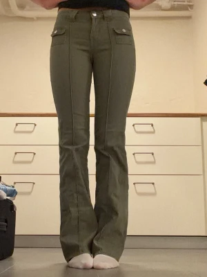 Olivgröna utsvängda jeans - Säljer ett par olivgröna jeans med utsvängda ben och låg midja. Jeansen har framfickor med lock och knapp, samt dekorativa sömmar längs benen. Jeansen är använda men fortfarande i ett bra sick💕💕