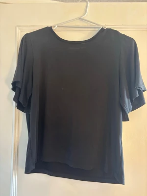Svart topp med fladdriga ärmar - En enkel svart topp med rund halsringning och fladdriga, korta ärmar. Toppen har en lös passform och är tillverkad i ett mjukt, tunt material som känns skönt mot huden. 