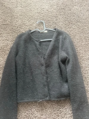 Mörkgrå fluffig kofta med knappar - Mysig mörkgrå kofta med fluffig textur och knäppning framtill. Koftan har lång ärm och en enkel, rak passform. Perfekt för lager på lager och ger en avslappnad look.