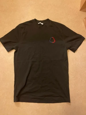 Svart Moncler t-shirt med logga - Säljer en svart t-shirt från Moncler med deras ikoniska logga i rött, blått och svart på bröstet. Klassisk passform och rund halsringning. Perfekt till jeans eller shorts för en clean och stilren look.