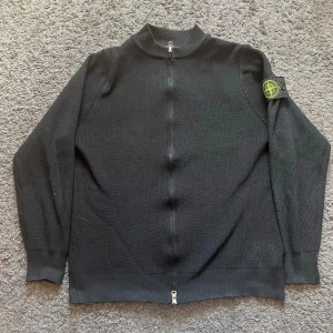 Svart stickad ziptröja Stone Island - Svart stickad ziptröja från Stone Island med ribbade muddar och hög krage. Tröjan har dragkedja framtill och den ikoniska Stone Island-loggan på vänster ärm. Perfekt för dig som gillar streetwear och snygga detaljer. Priset kan diskuteras 