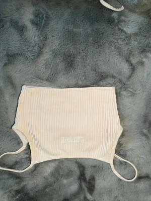 Beige ribbad bandeau från Lounge - Säljer en beige ribbad bandeau-topp från Lounge Toppen är axelbandslös med smala band och har en broderad logga framtill. Perfekt för sommardagar eller som layering-piece. Supersnygg och enkel att matcha med olika outfits!