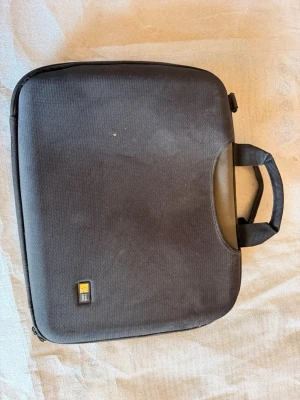 Svart laptopväska med handtag - Säljer en svart datorväska med dubbla handtag och slitstarkt tygmaterial. Väskan har en enkel design med diskret logga på framsidan och praktiskt band på baksidan för att fästa på resväska. Perfekt för att bära med sig laptopen till skolan eller jobbet.