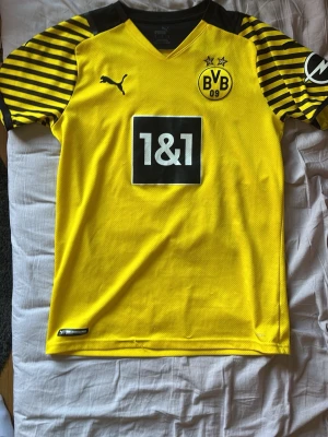 Borussia Dortmund matchtröja Puma - Säljer en gul och svart Borussia Dortmund matchtröja från Puma med klubbmärke, sponsortryck och två stjärnor på bröstet. Tröjan har svarta och gula ränder på ärmarna och texten 'DORTMUND' på ryggen. Perfekt för fotbollsfans!