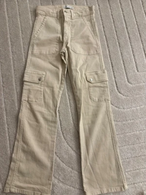 Beige cargobyxor med fickor - Snygga beige cargobyxor med raka ben och stora fickor på sidorna med lock och knapp. Byxorna har även två framfickor och är tillverkade i ett slitstarkt bomullstyg. Perfekta för en avslappnad och trendig look.