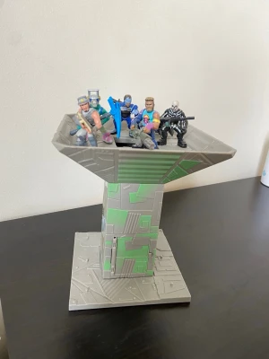 Fortnite leksakstorn med figurer - Säljer ett Fortnite-inspirerat leksakstorn i grått och grönt med flera actionfigurer på toppen. Tornet har en lucka med ett vapen gömt inuti. Perfekt för dig som gillar gaming och samlarprylar. Figurerna är färgglada och detaljerade.