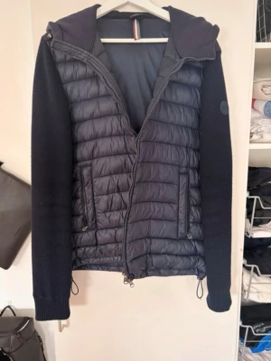 Colmar cardigan - Snygg svart cardigan med stickade ärmar och quiltad front. Jackan har dragkedja, två fickor fram och en vadderad huva. Materialmix med både vadderad och stickad känsla ger en cool look. Perfekt för kyliga dagar och enkel att matcha till olika outfits.