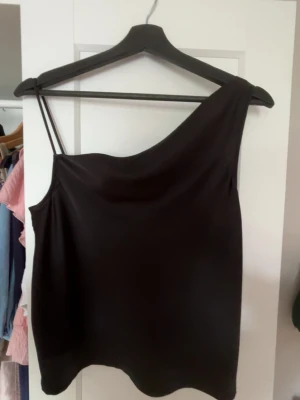 Svart oneshoulder topp - Snygg svart topp med oneshoulder-design och smalt axelband på ena sidan. Perfekt för dig som vill ha en stilren och trendig look. Materialet känns mjukt och har ett lätt glansigt intryck.