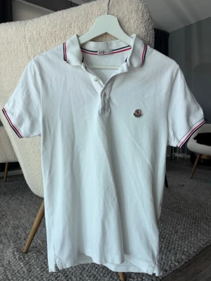 Moncler piké - Klassisk vit pikétröja från moncler med korta ärmar och krage. Tröjan har rödblå ränder på krage och ärmslut samt en liten broderad logga på bröstet. Tillverkad i mjuk bomull och har knappar vid halsen för en sportig look.