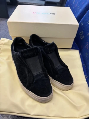 Svarta slip-on sneakers Axel Arigato - Stilrena svarta slip-on sneakers från Axel Arigato i sammet med bred resår över vristen. Skorna har en vit, tjock sula och minimalistisk design utan snörning. Perfekta för en clean och modern look.