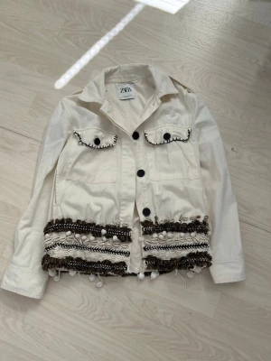 Vit jeansjacka med detaljer från Zara - Snygg vit jeansjacka från Zara med dekorativa svarta och bruna broderier samt bollar längs nederkanten. Två bröstfickor med lock och svarta knappar framtill. Perfekt för dig som vill sticka ut med coola detaljer.