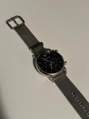 Emporio Armani silverfärgad klocka - Stilren klocka från Emporio Armani med svart urtavla och mesh-armband i silverfärgat metall. Klockan har kronograf och minimalistisk design som passar till alla outfits. Perfekt accessoar för dig som gillar klassisk men modern stil.