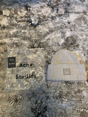 mössa Acne Studios - Säljer en ljusgrå ribbstickad mössa från Acne Studios med uppvikt kant och det klassiska Face-logotypen framtill. Mössan är tillverkad i mjukt material och har en stilren, minimalistisk design som passar perfekt till höst och vinter.