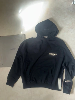 Svart Essentials Fear of God hoodie - Snygg svart hoodie från Essentials Fear of God med vit logga på bröstet och ärmen. Klassisk känguruficka framtill och stor huva. Tillverkad i mjukt bomullsmaterial som känns riktigt skönt mot huden. Perfekt för dig som gillar streetwear och enkel stil. Ej använd storlek L