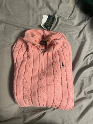 Rosa kabelstickad tröja Polo Ralph Lauren - Kabelstickad rosa tröja från Polo Ralph Lauren med halv dragkedja och hög krage. Tröjan har det klassiska broderade logot på bröstet i mörkblått och ribbade muddar vid ärmslut och nederkant. Perfekt för dig som gillar preppy stil. sitter som Xl