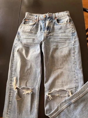 Gina Tricot ripped jeans 🤍 | ljusblå - Säljer ett par ljusblå mom jeans med slitna detaljer och stora hål vid knäna. Jeansen har hög midja, klassisk femficksmodell och raka ben. Perfekta för en avslappnad och trendig look.