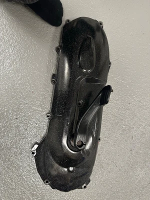 Svart variatorkåpa till moped - Säljer en svart variatorkåpa till moped, tillverkad i metall. Kåpan har flera skruvhål runt kanterna och en pedalarm monterad på ena sidan. Passar till dig som behöver reservdel till din scooter eller moped.