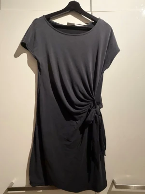 Svart kort klänning med knyt - En svart kort klänning med rund halsringning och korta ärmar. Klänningen har en snygg knytning i sidan som ger en draperad effekt. Perfekt för dig som gillar enkel men stilren design.