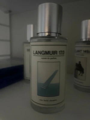 Langmuir 1720 Eau de Parfum - En stilren parfymflaska med silverfärgat lock och minimalistisk etikett. Flaskan är genomskinlig och etiketten har en modern design med blått motiv. Doften heter Langmuir 1720 och är en eau de parfum.