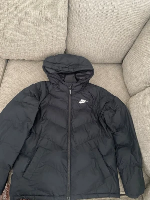 Nike jacka  - Snygg svart pufferjacka från Nike med huva och dragkedja framtill. Jackan har en klassisk vadderad look och Nike-logga på bröstet. Perfekt för kalla dagar och riktigt skön att bära. Materialet är slitstarkt och håller värmen bra, jackan passar som S