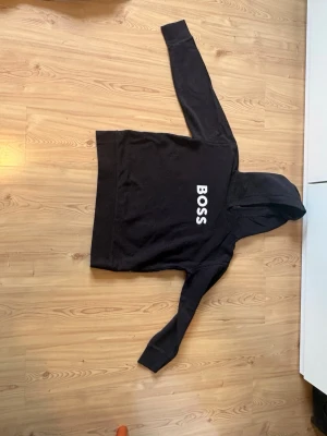 Svart hoodie med BOSS-tryck - Säljer en svart hoodie med stor vit BOSS-text på bröstet. Klassisk modell med huva och långa ärmar. Hoodien har en avslappnad passform och är tillverkad i mjukt material, perfekt för en chill och stilren look.