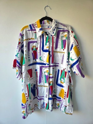 Färgglad vintage skjorta Polarn & Pyret - Unik vintage skjorta från Polarn & Pyret med färgglatt abstrakt mönster i lila, blått, grönt, rött och gult på vit botten. Skjortan har korta ärmar, klassisk krage och knäppning framtill. Perfekt för dig som gillar retrostil och statement-plagg.
