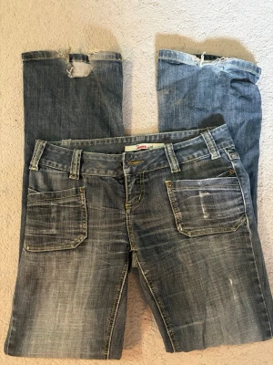 Blå slitna jeans med raka ben - Ett par blå jeans med raka ben och slitna detaljer, inklusive hål och fransar vid bensluten. Jeansen har stora framfickor och bakfickor med lock. Klassisk femficksmodell i denim med tvättad look och kontrastsömmar.