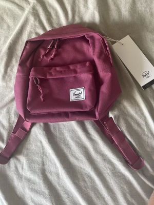 Vinröd ryggsäck från Herschel - Snygg vinröd ryggsäck från Herschel med klassisk design. Väskan har ett stort huvudfack, en mindre ytterficka med dragkedja och justerbara axelband. Tillverkad i slitstark canvas och har randigt innerfoder. Perfekt för skola eller cityhäng.