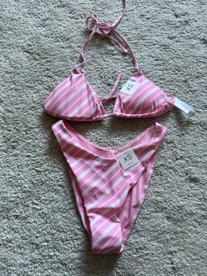 Rosa och vit randig bikini XS - Snygg bikini i triangelformad modell med rosa och vita ränder. Överdelen har knyt i nacken och ryggen, och underdelen har hög midja. Perfekt för strandhäng och poolparty.