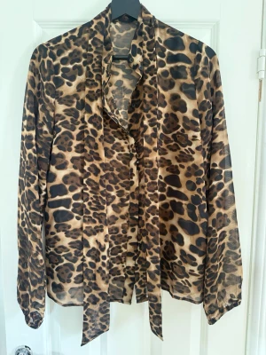 Leopardmönstrad blus från Gestuz - Snygg leopardmönstrad blus från Gestuz i tunnt, glansigt material. Blusen har långa ärmar och en dekorativ knytband vid halsen som ger en cool och trendig look. Perfekt för dig som vill sticka ut med ett djurmönster. Även lite stor i storleken.