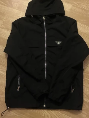 Prada windbreaker - Snygg svart vindjacka från Prada med huva och dragkedja framtill. Jackan har två fickor med dragkedja och det klassiska Prada-emblemet på bröstet. Tillverkad i ett lätt och tåligt material som passar perfekt för blåsiga dagar.