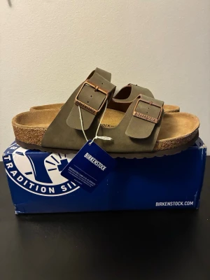 Birkenstock Arizona storlek 43 - Säljer ett par Birkenstock Arizona sandaler i olivgrönt mockaläder med två breda remmar och justerbara metallspännen. Fotbädden är i mjukt naturfärgat läder och sulan är i kork för extra komfort. Perfekta för en avslappnad och trendig stil.