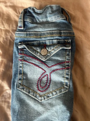 Blå bootcut jeans med hål i knäna och röda detaljer - Blåa bootcut jeans från Gina tricot med hål vid knäna, knappar på fickorna och röda detaljer.