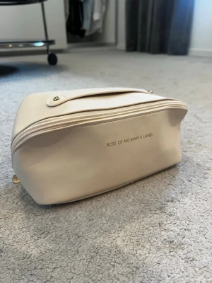 Beige necessär med dragkedja - Beige necessär med  ett praktiskt handtag upptill och dragkedja. Invändigt finns två stora fack och elastiska fickor för enkel organisering. Tillverkad i ett slätt, lättskött material.